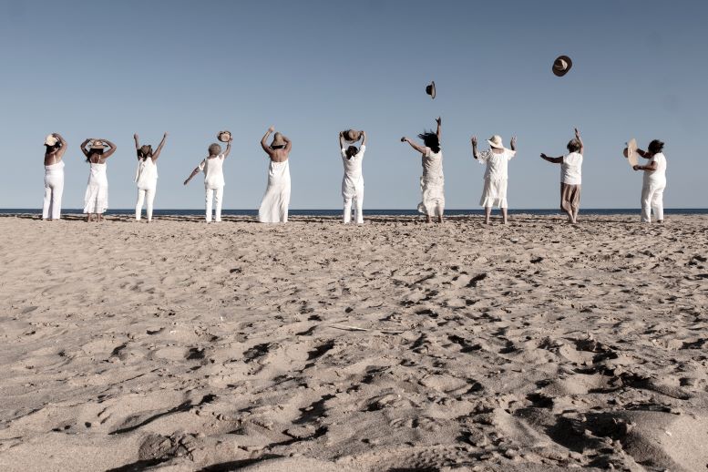 Várias mulheres, vestidas de branco e de costas, na praia. Autora da fotografia é Maria Manuel Pinto.