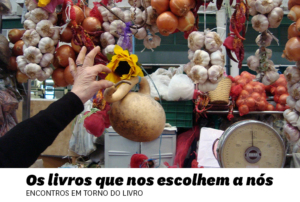 Fotografia com uma mão a escolher um girassol no meio de alhos e cebolas, num ambiente de mercado.