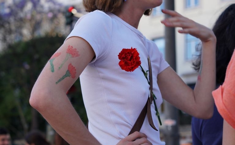 Imagem ilustrativa do evento. Fotografia com uma mulher de camisola branca com cravos vermelhos. A fotografia tem Direitos Reservados.