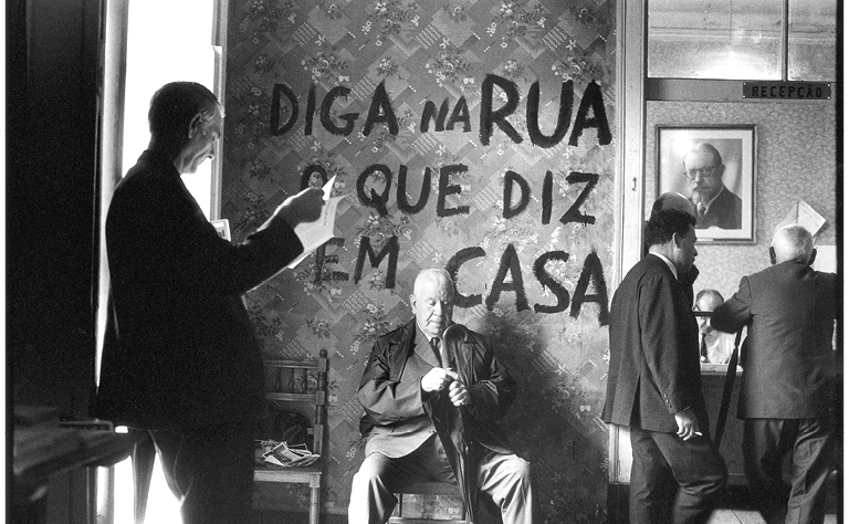 Fotografia de Carlos Gil, a preto e branco, onde se lê a frase "Diga na rua o que se diz em casa".