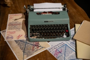 Imagem com uma máquina de escrever sobre vários textos impressos e censurados. Autoria da fotografia de Mariano Teixeira.