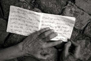 Fotografia a preto e branco de duas mãos de uma pessoa a escrever num guardanapo de papel. Foto da autoria de Valentina Parravicini.